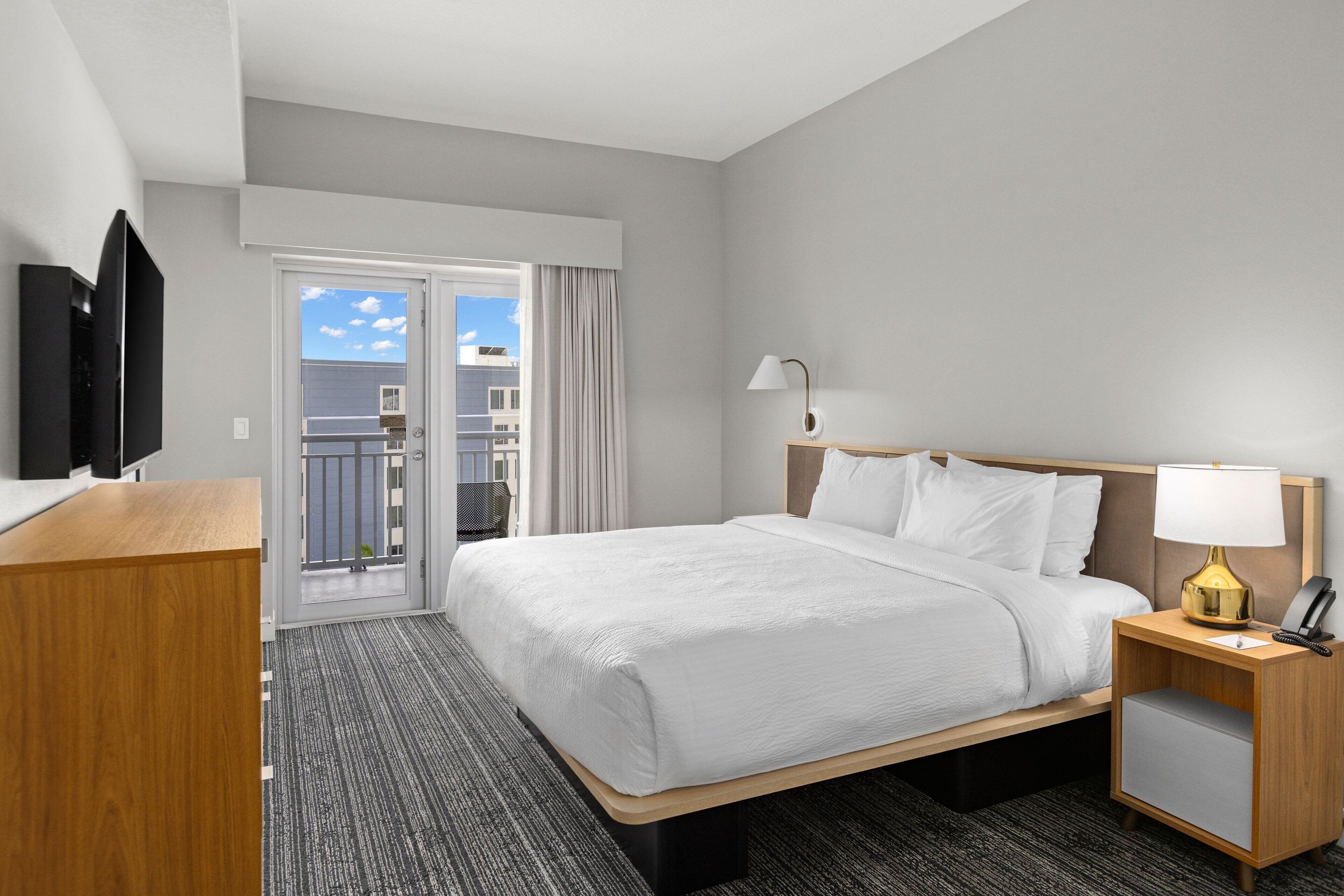 A hotel room with a large bed, a wooden nightstand with a lamp, a desk with a television, and a sliding door leading to a balcony.