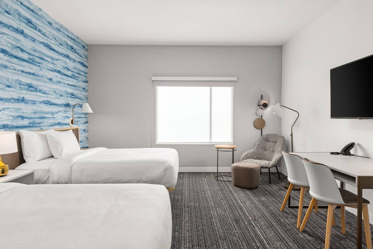 Ein Hotelzimmer mit zwei Betten, einem Schreibtisch, einem Sessel, einem kleinen runden Tisch, einer Lampe und einem an der Wand montierten Fernseher.