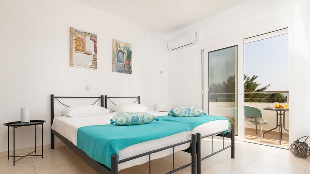 A bedroom with a bed, a small bedside table, a balcony with a table and chairs, and two wall-mounted artworks. - Lungo Mare (Rhodes)