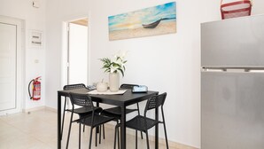 A dining area with a table, chairs, a vase with flowers, a painting of a boat on the beach, and a refrigerator. - Lungo Mare (Rhodes)