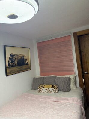 A bedroom with a bed, pillows, a blanket, a framed picture, a window with blinds, and a door.