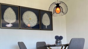 Een eetgedeelte met een moderne tafel, stoelen, een kroonluchter en drie kunstwerken in lijsten.