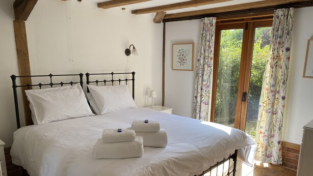 Una cama bien hecha con sábanas blancas y dos toallas dobladas, suelo de madera, mesita de noche blanca, lámpara de pared y ventana con cortinas florales.