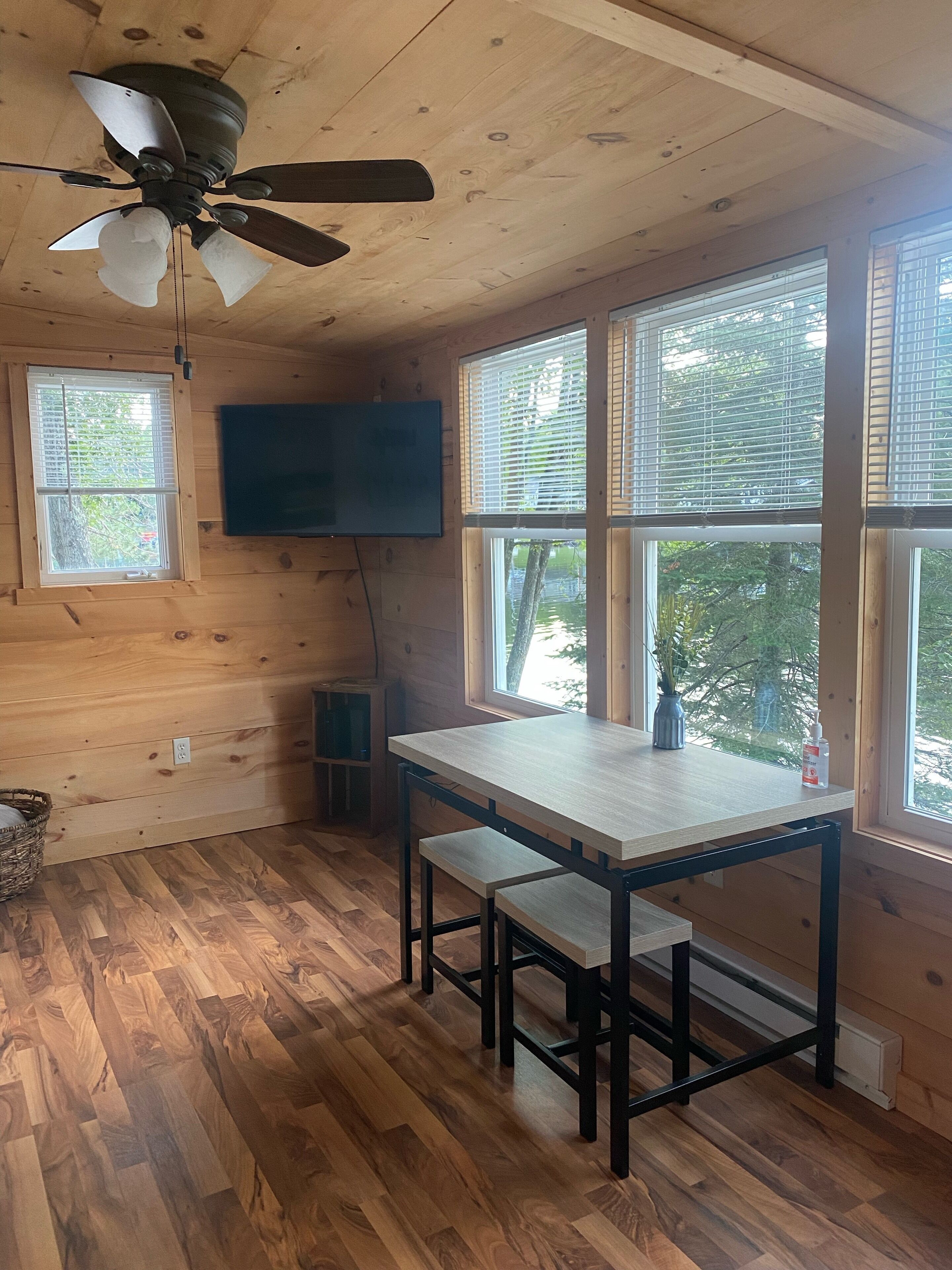 A wooden cabin interior with a dining table, two benches, a ceiling fan, a television, and large windows.