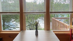 Una mesa de madera para comer con bancos, tres ventanas con persianas y vista a una terraza y árboles.