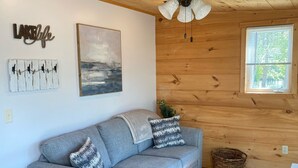 Una habitación acogedora con un sofá gris, una pequeña mesa redonda, un ventilador de techo, paredes de madera, una ventana y una obra de arte colgada en la pared.
