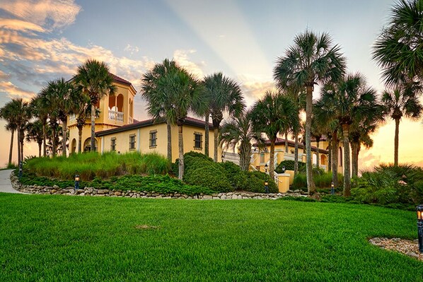 A large, yellow building with a balcony, surrounded by palm trees and a well-maintained lawn.