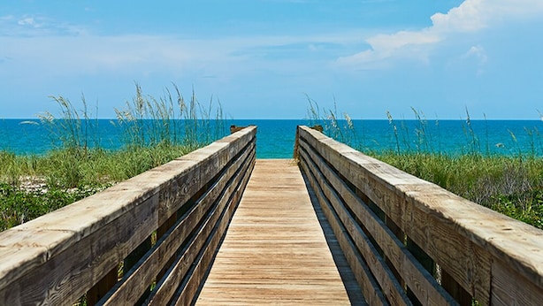 Een houten looppad dat naar een strand leidt, met helderblauw water en begroeiing aan weerszijden.