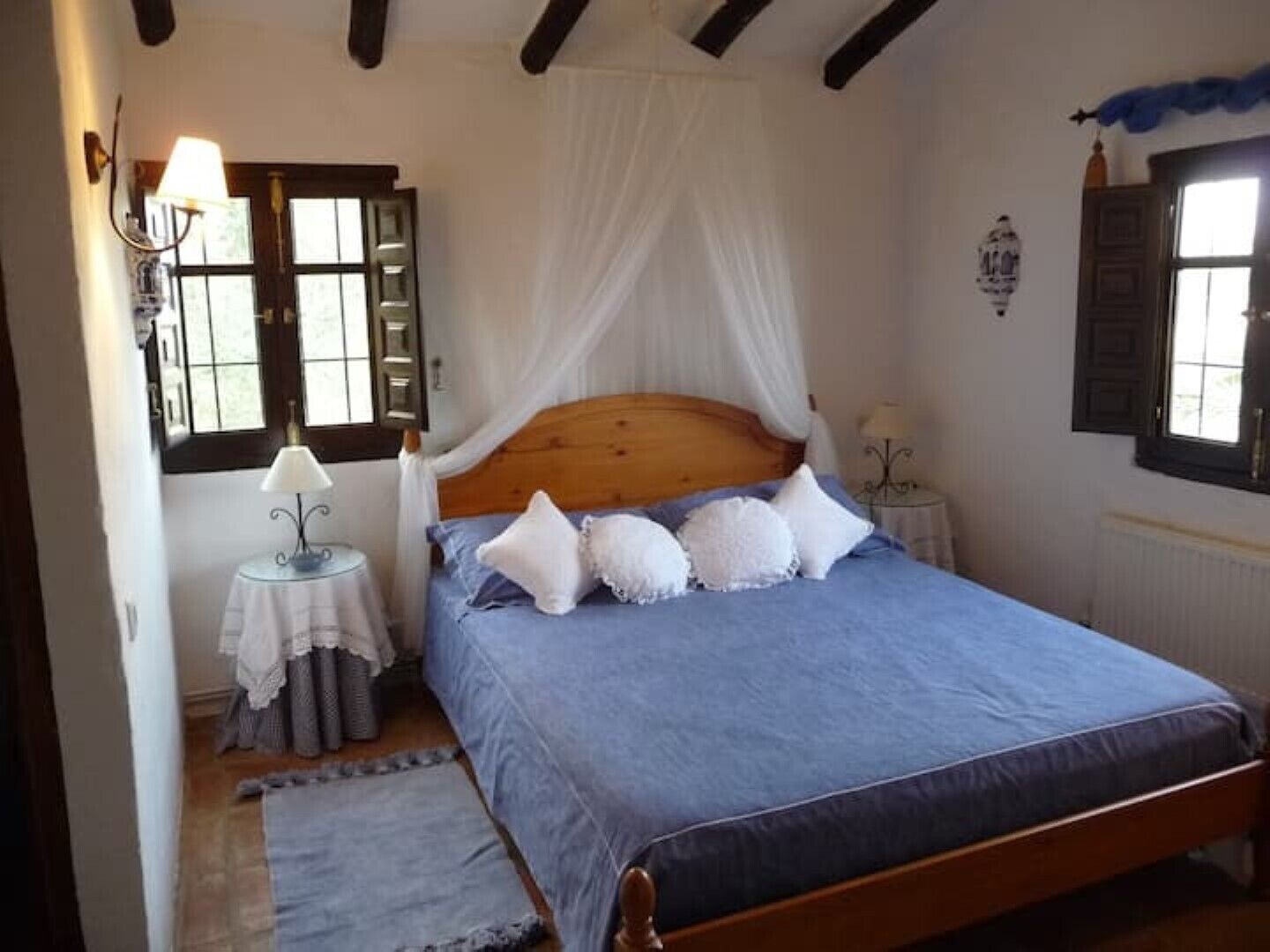 A bedroom with a wooden bed, a canopy, a small bedside table with a lamp, and a window with curtains.
