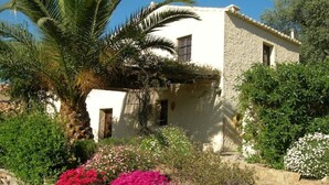 A rustic stone building with a white facade, surrounded by lush greenery and flowering plants.