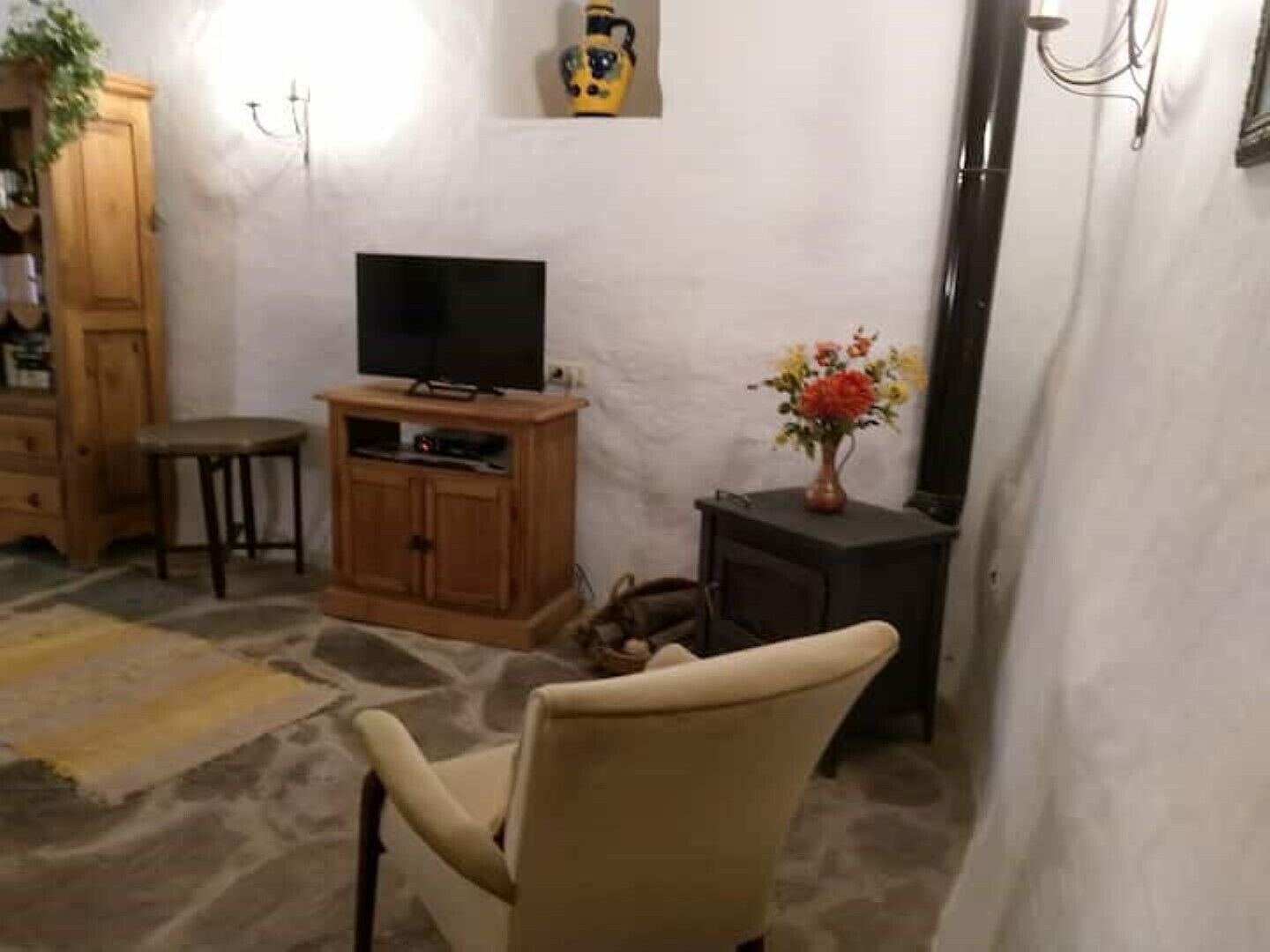 Een woonkamer met een televisie op een houten kast, een stenen vloer, een beige fauteuil en een tafeltje met een bloemenvaas.