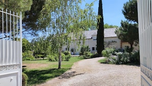 Une allée de gravier mène à un bâtiment en pierre, agrémenté d’un jardin et d’un grand arbre.