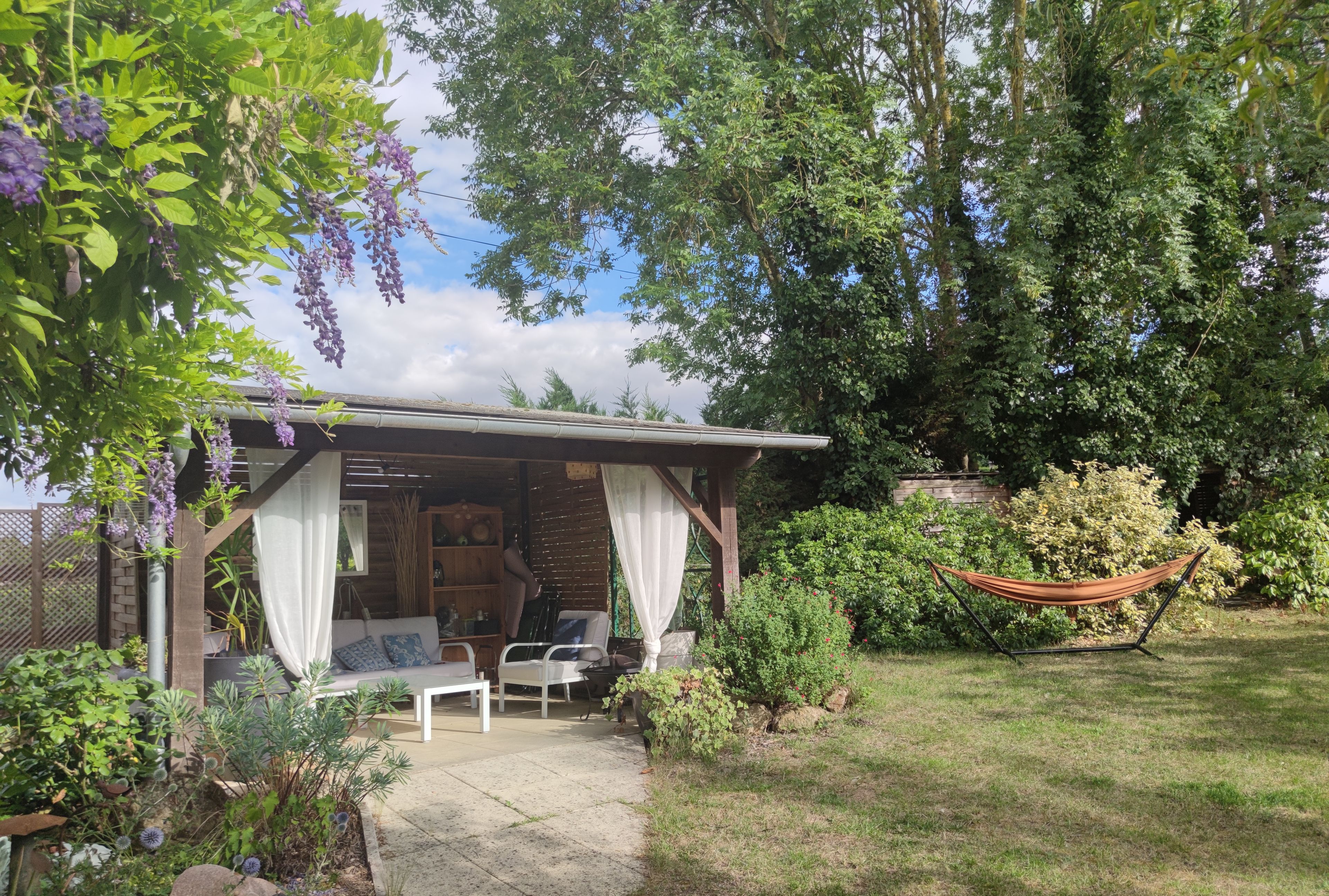 Een overdekt terras met een tafel en stoelen, omgeven door weelderig groen en een hangmat.