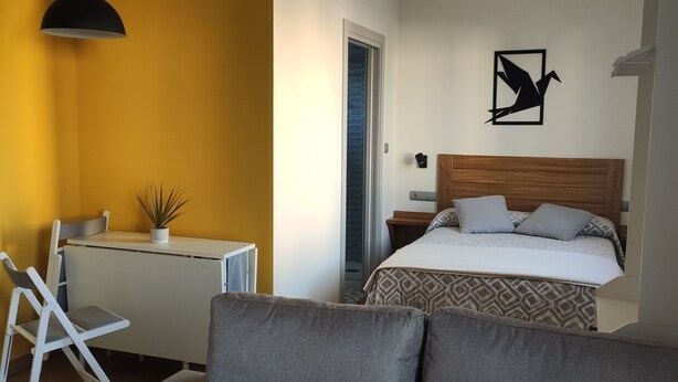 Un dormitorio moderno con un sofá gris, una mesita con una planta, una cama con ropa de cama estampada y una obra de arte en la pared.