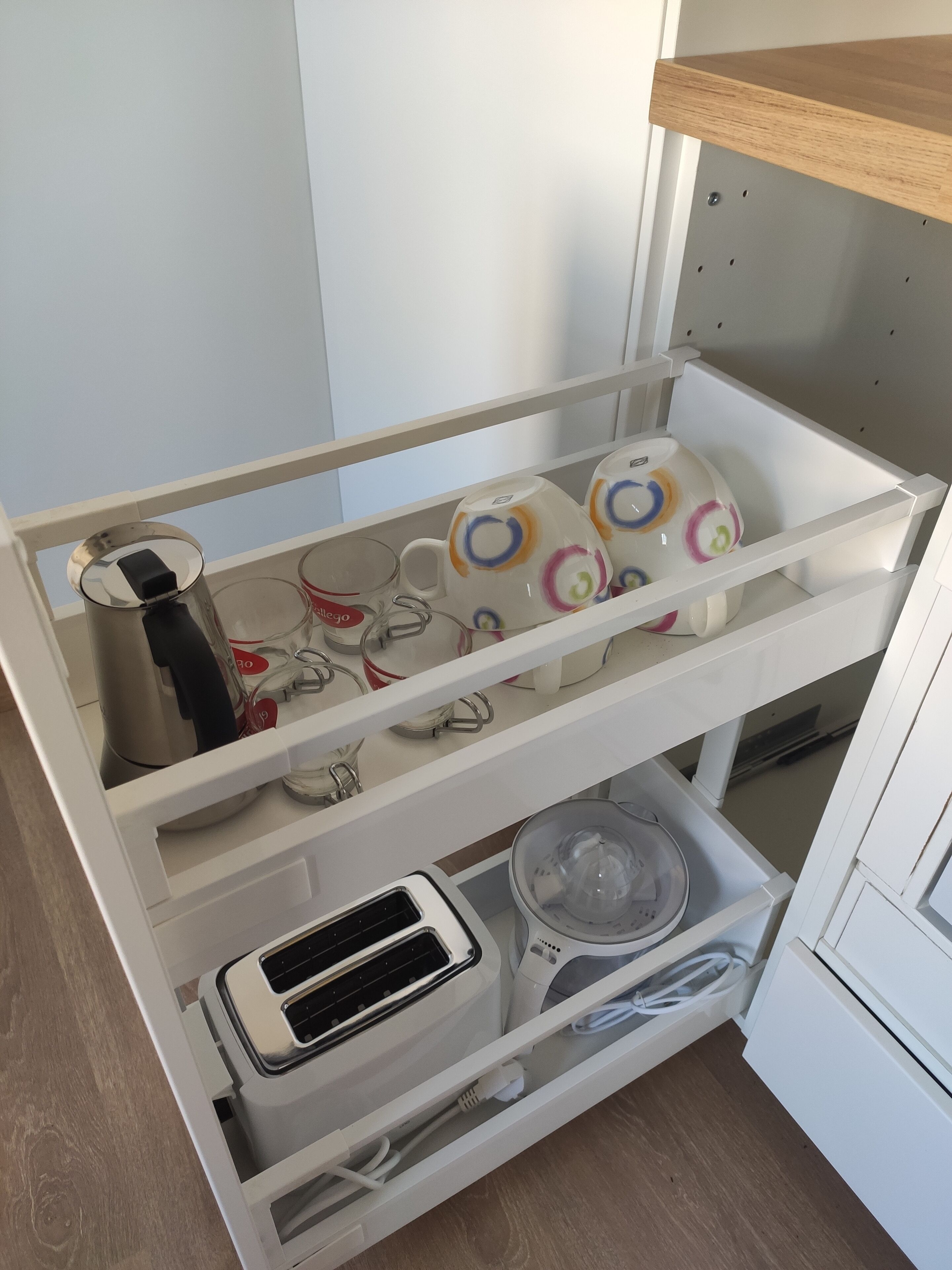 Carrito de cocina blanco con estantes que contienen tazas, una tetera, un tostador y una licuadora.