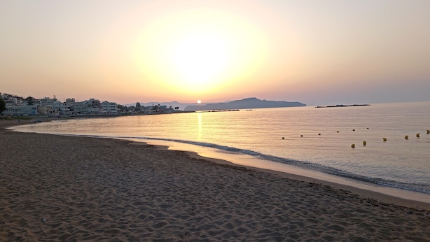 Una platja amb un capvespre, mar tranquil·la i edificis al fons.