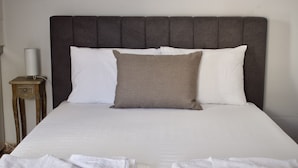 Un llit ben fet amb un capçal fosc, almoades blanques i una almoadeta decorativa marró. Hi ha una petita taula de nit de fusta amb una làmpada al costat del llit.