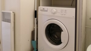 Kompakti pyykinhuoltotila, jossa on etuaukosta täytettävä pesukone, seinälle asennettu lämmitin ja seinää vasten nojaava harja.