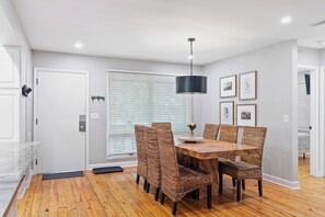 A dining area with a wooden table and wicker chairs, a black pendant light, and framed pictures on the wall.