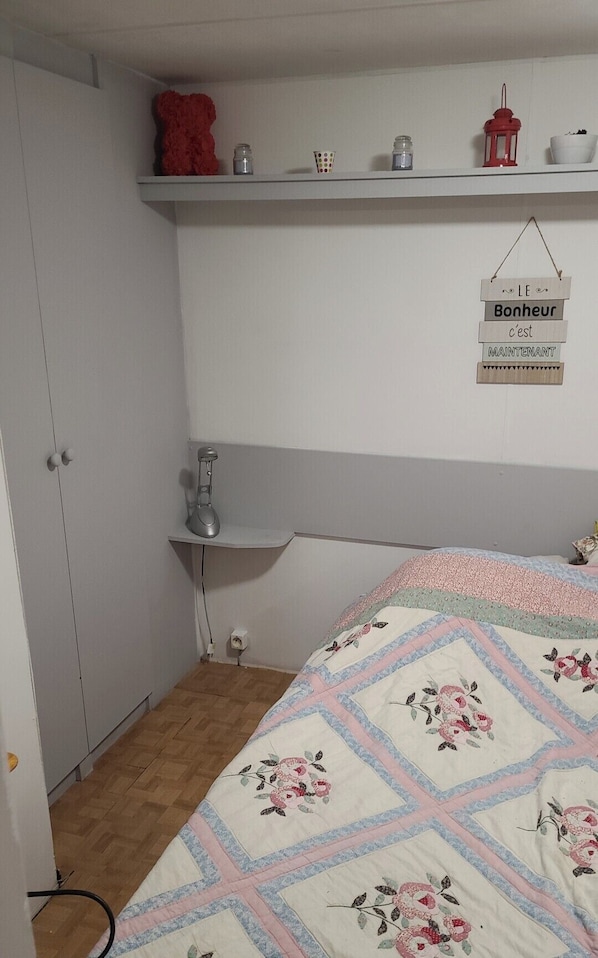A bedroom with a bed, a shelf with decorative items, a hanging sign, and a desk with a lamp. - Mobile home (Mimizan)