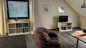 Un salon chaleureux avec une grande fenêtre, un téléviseur, un fauteuil à motifs et une petite table sur laquelle est posée une plante.
