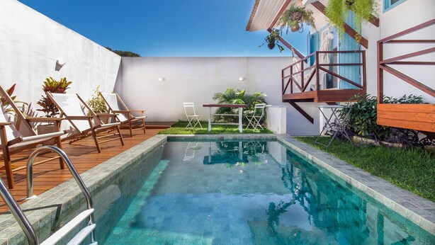 Una piscina con sillones y mesas de madera, rodeada de plantas en macetas y un muelle de madera.