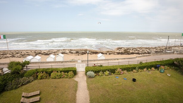 Blick auf einen Strand mit einem Pfad, der zu Strandkörben und einem Rettungsturm führt, sowie eine Person beim Kitesurfen.