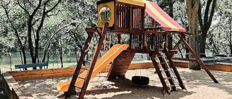 Une balançoire en bois avec un toboggan rouge et jaune, entourée de sable et d’arbres.