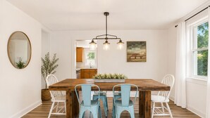 A dining room with a wooden table, four chairs, a hanging light fixture, and a painting on the wall.