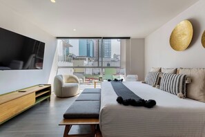 A modern hotel room with a large bed, a sofa, a TV, and a view of the cityscape through the window.
