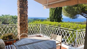 A terrace with a table and chairs, stone pillar, white lattice railing, and a view of the sea.