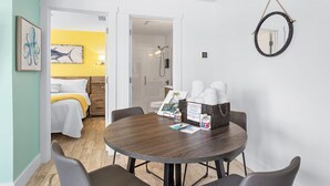 A hotel room with a round dining table, a bed with a yellow blanket, a shower, and a wall-mounted mirror. - Deluxe King Suite w/Deck (Saint James City)