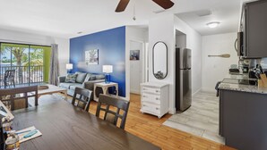 A modern living room with a ceiling fan, a dining area with a wooden table and chairs, a kitchen with a refrigerator and a microwave, and a blue accent wall. - Deluxe King Suite w/Deck (Saint James City)