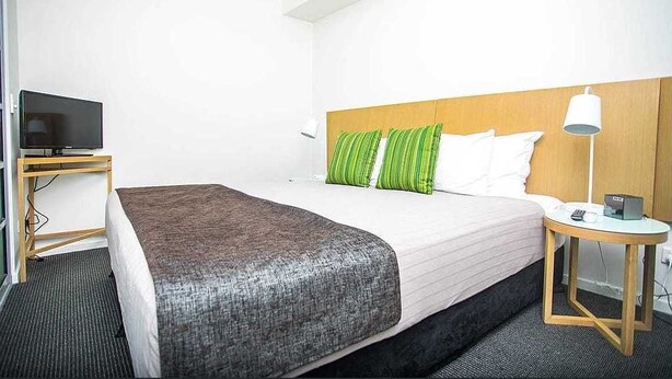 A hotel room with a bed, a TV on a stand, a bedside table with a lamp, and a small table with a phone and a box.