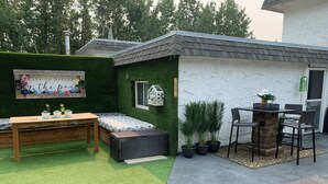Zona de pati exterior petita amb taula de fusta, una banca i un llit petit amb paret verda i gespa artificial.
