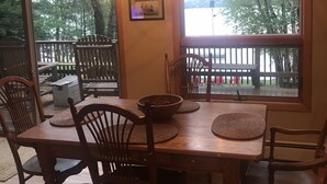A dining area with a wooden table and chairs, a hanging light fixture, and a large window with a view of trees and a dock. - Lake front beauty (Hillman)
