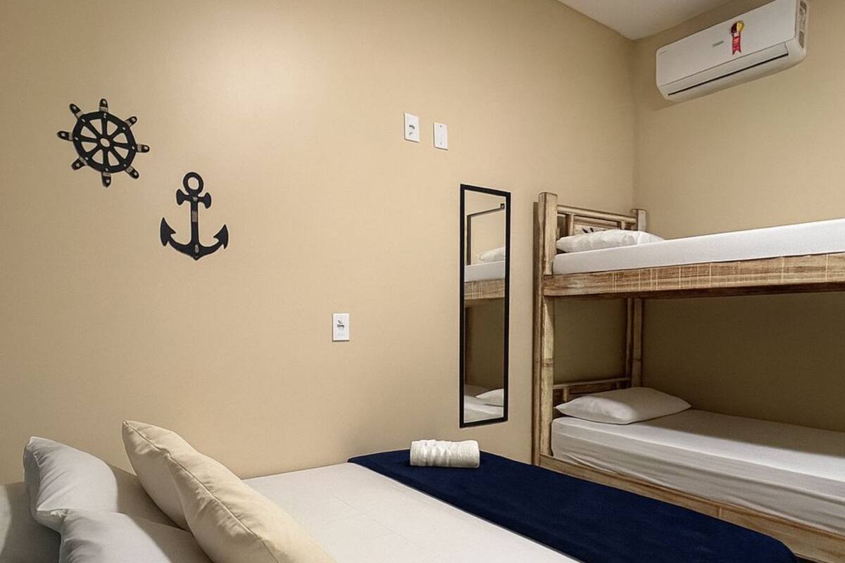 A single bed with a blue blanket and two pillows, a bunk bed with white bedding, a wall-mounted air conditioner, and two wall decorations (a ship's wheel and an anchor).