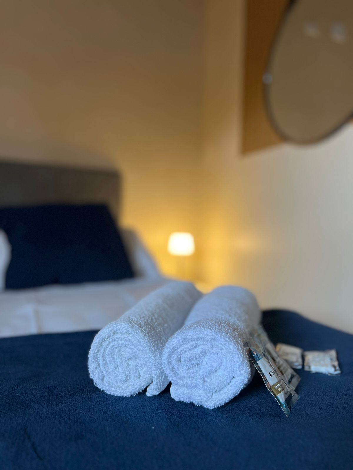 A bed with a dark blue bedspread, two rolled white towels, and a packet of toiletries.