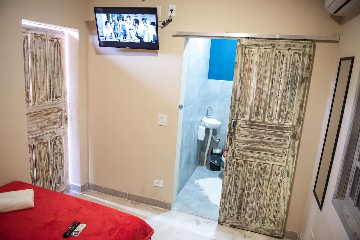 A room with a red bed, a television mounted on the wall, a shower with a blue curtain, and a patterned door.