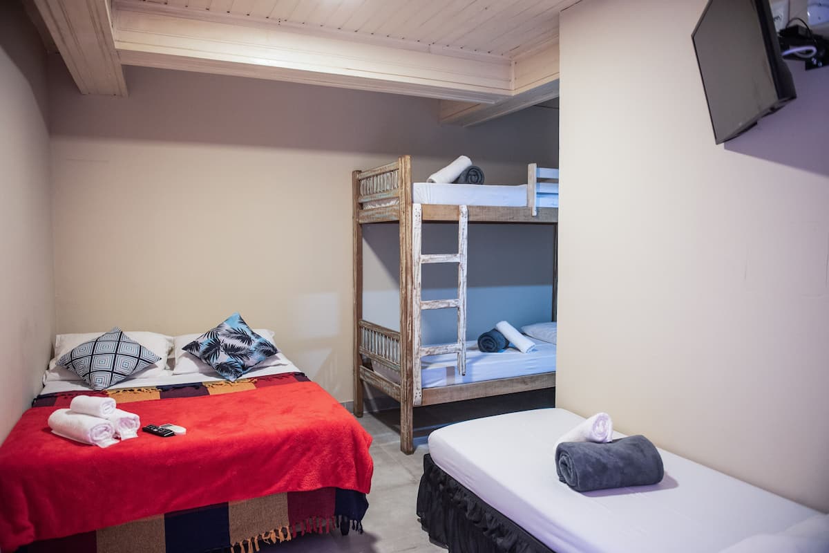 A room with two beds, one with a red blanket and the other with a grey blanket, a wooden bunk bed, and a television mounted on the wall.