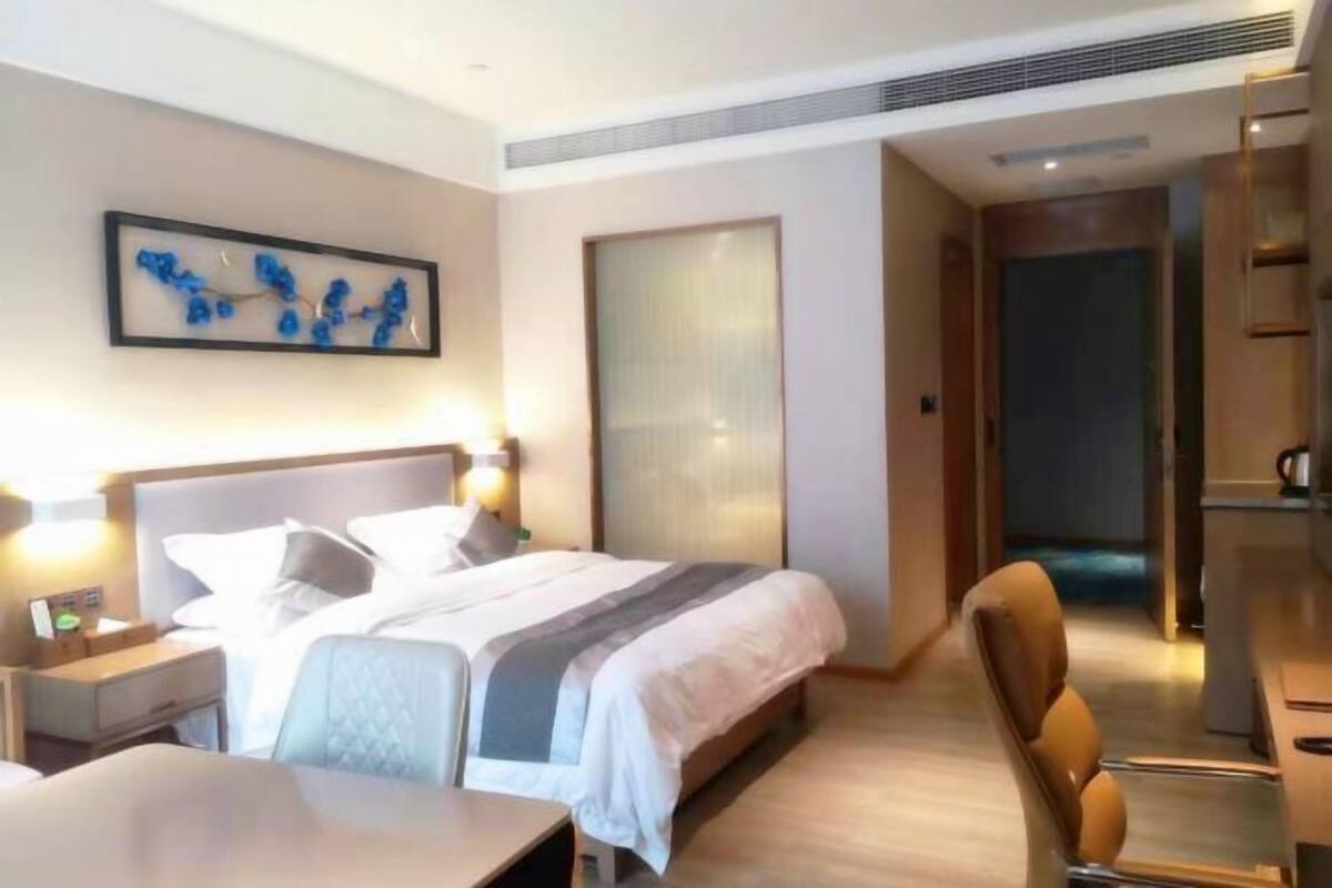 A hotel room with a bed, bedside table, chair, and a desk. There is a painting on the wall and a door leading to another room.