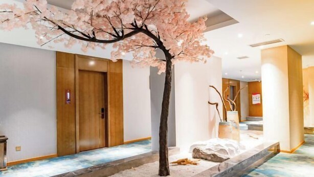 A hotel lobby with a central artificial cherry blossom tree, a stone pathway, and a wooden door in the background. - Xingzhike Hotel (Wuchuan)