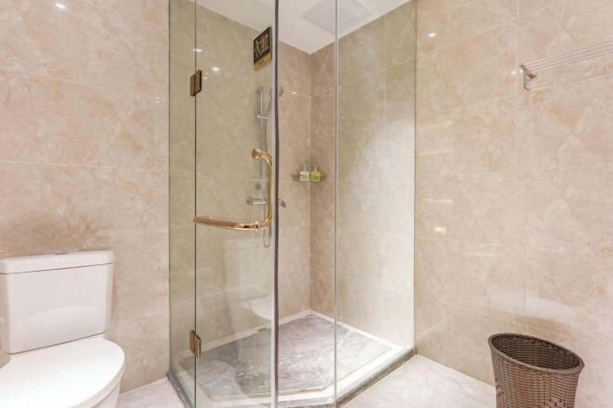Ducha con mampara de vidrio, inodoro blanco y una cesta de mimbre en un baño con paredes de mármol.