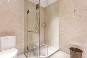 A glass-enclosed shower with a white toilet and a wicker basket in a bathroom with marble walls.