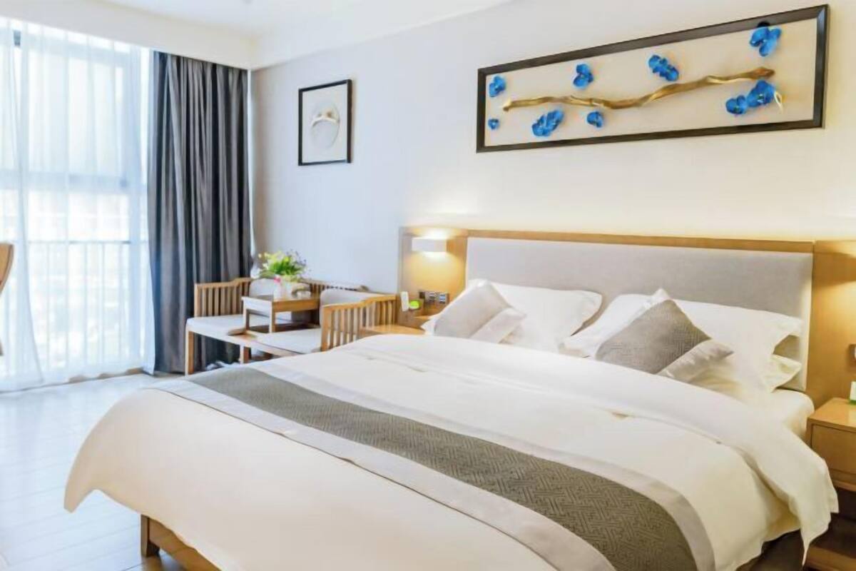 A hotel room with a large bed, a bedside table, a desk with a chair, and a window with curtains.