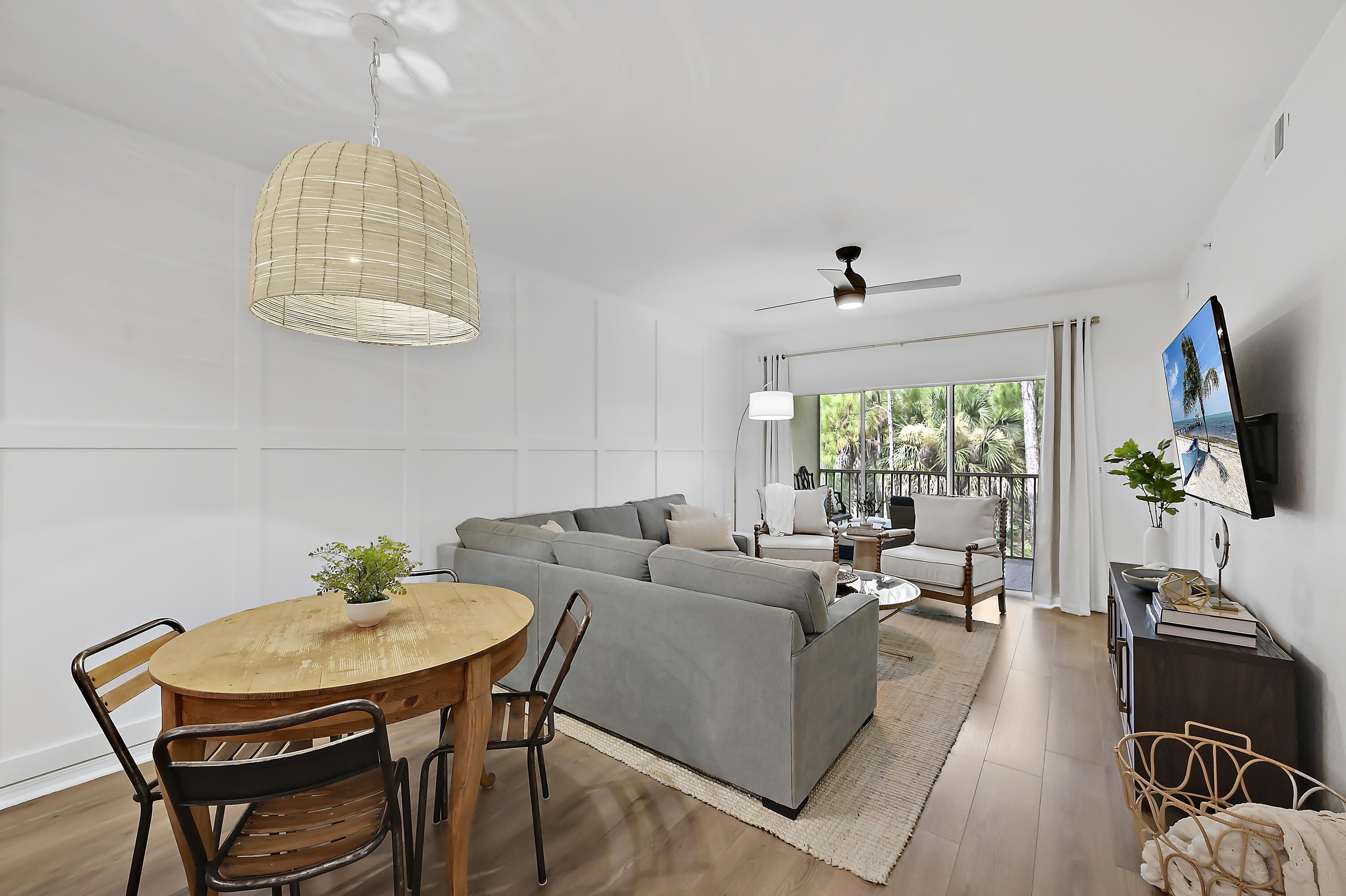 Uma sala de estar moderna com sofá seccional cinza, mesa de jantar de madeira e cadeiras. Há uma janela grande com vista para palmeiras.