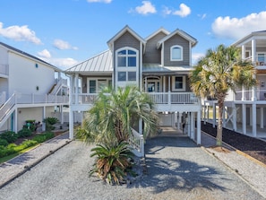 A two-story beachfront property with a covered porch, a staircase, and a gravel driveway.