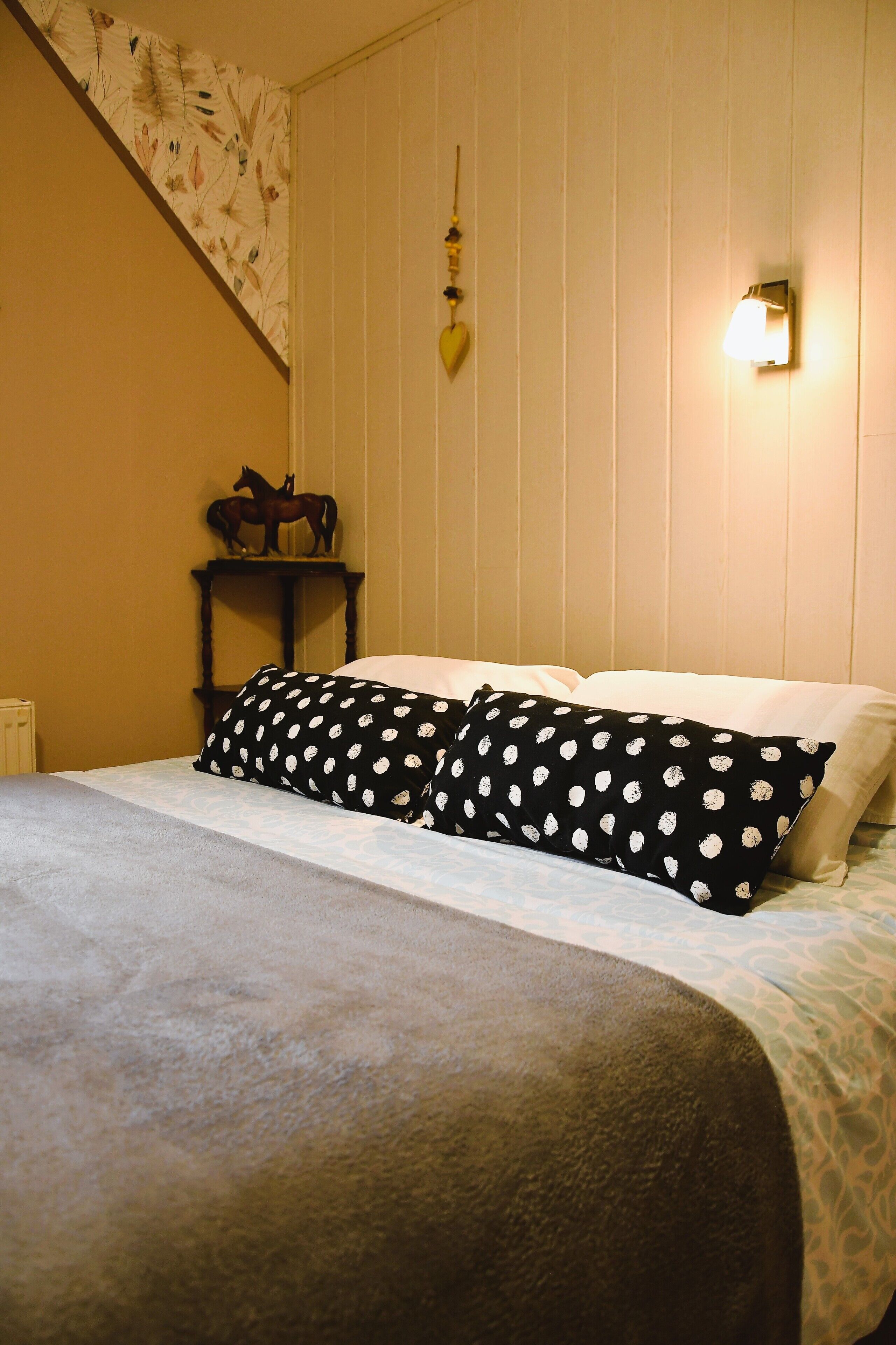 A bedroom with a bed, a nightstand with a horse figurine, a wall-mounted light, and a staircase with floral wallpaper.