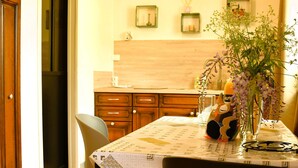A dining area with a table set for two, a vase with flowers, and a kitchen in the background.