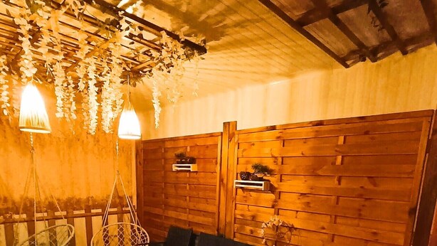 A hot tub with a control panel, surrounded by wooden walls and hanging lights. There are two chairs and a small table with a basket.